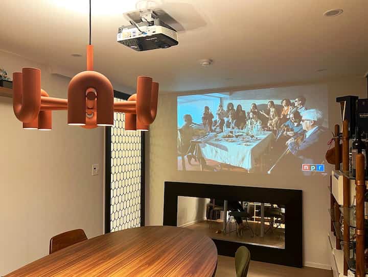 Natural tone dining room featuring a large oval table, chandelier, and ceiling-mounted projector creating a home cinema atmosphere