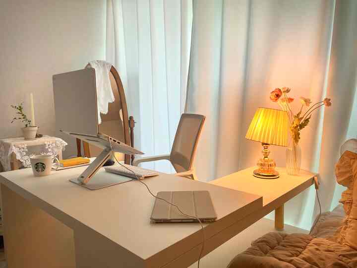 白とベージュのトーンのホームオフィス、白いデスクと椅子にノートパソコンスタンド、暖かい照明と花瓶が配置された印象的な空間