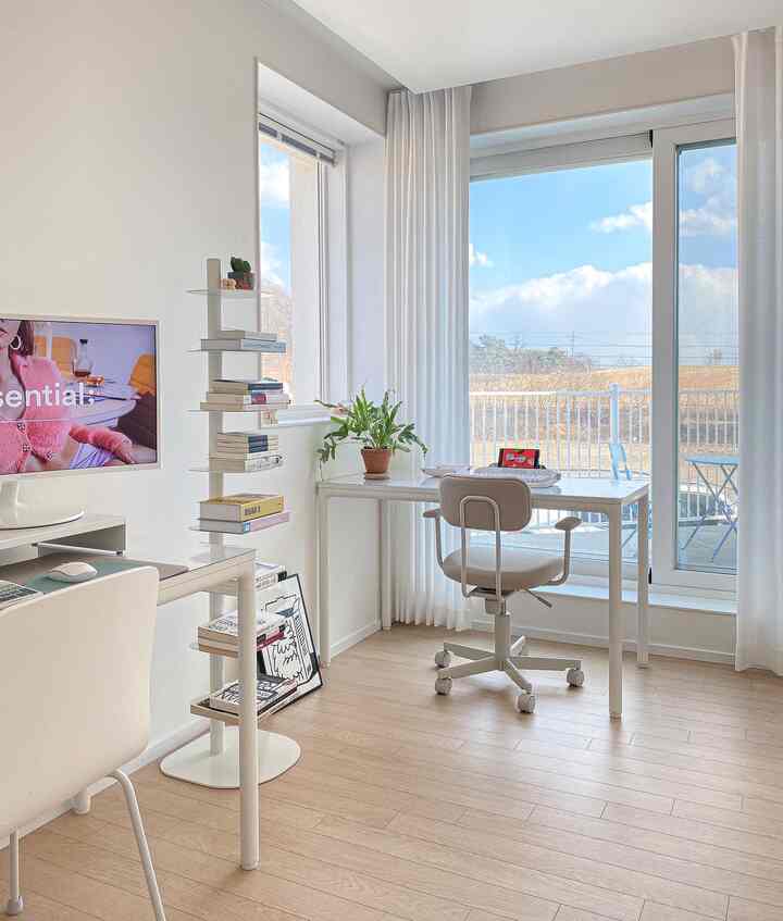 Bright and clean white and beige toned home office featuring desks, office chair, bookshelf, and curtains with natural light
