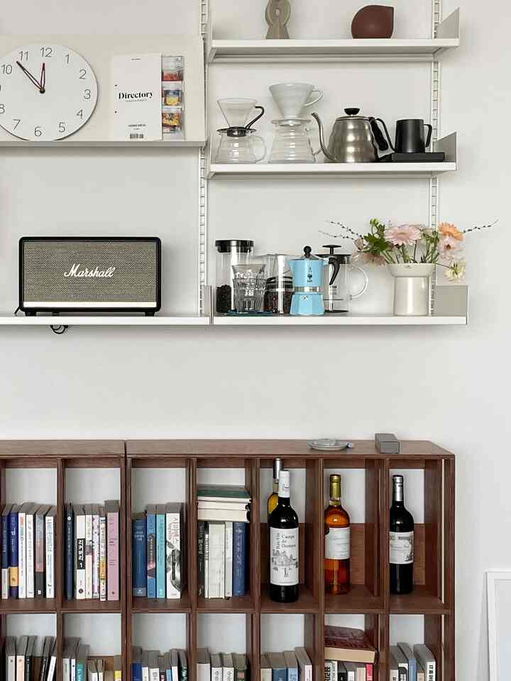 A white-walled home cafe space featuring white shelves and a brown wooden bookshelf, decorated with coffee equipment and a flower vase creating a neat, cozy atmosphere.