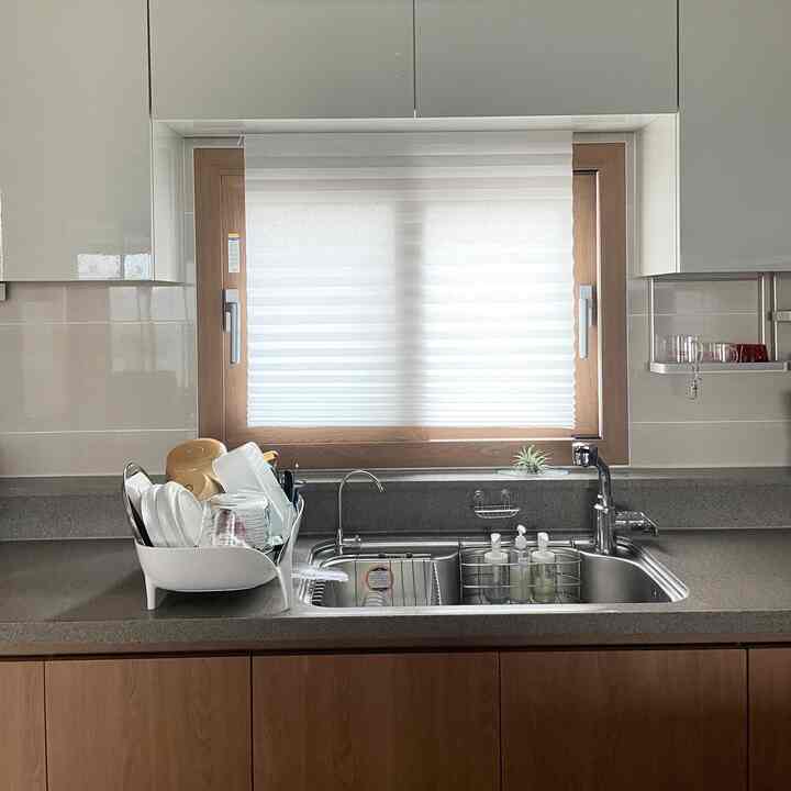 Clean kitchen with white and wood tones, featuring blinds on the window and a dish drying rack on the countertop for practical use