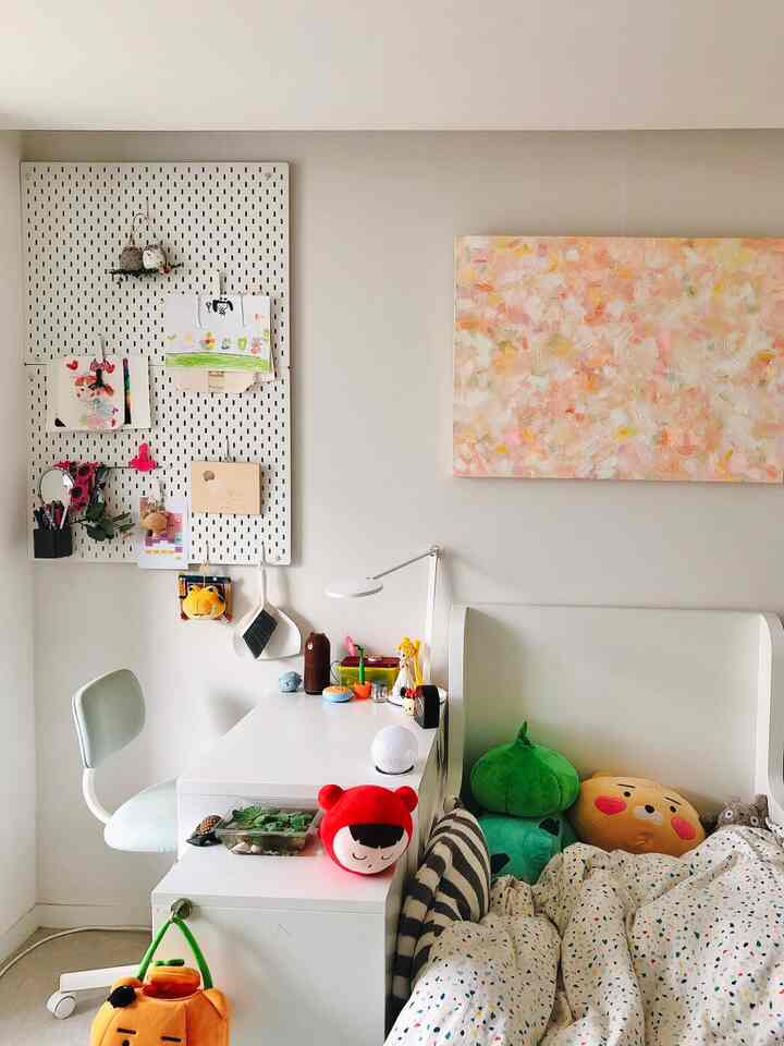 White and natural tone home office featuring a desk, children's drawings on pegboard, toys on bed in a simple room