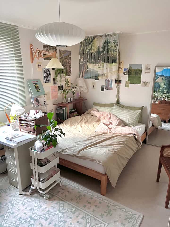 A beige and natural wood-toned studio bedroom featuring a wooden bed frame, IKEA desk, plants, and fabric posters creating a cozy atmosphere