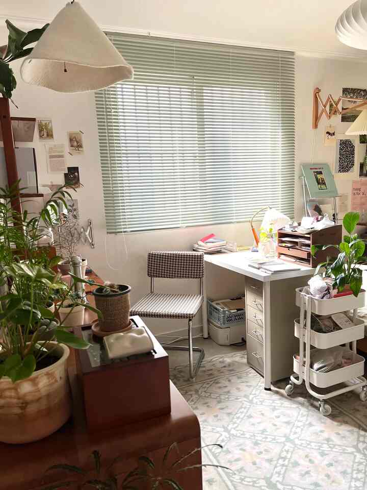 Studio apartment home office with light green blinds, wood furniture, and numerous plants creating a cozy atmosphere