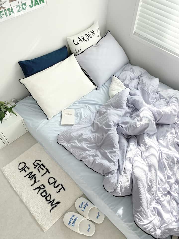 Light blue and white toned small bedroom featuring tidy bedding, cushions, and natural light from window blinds