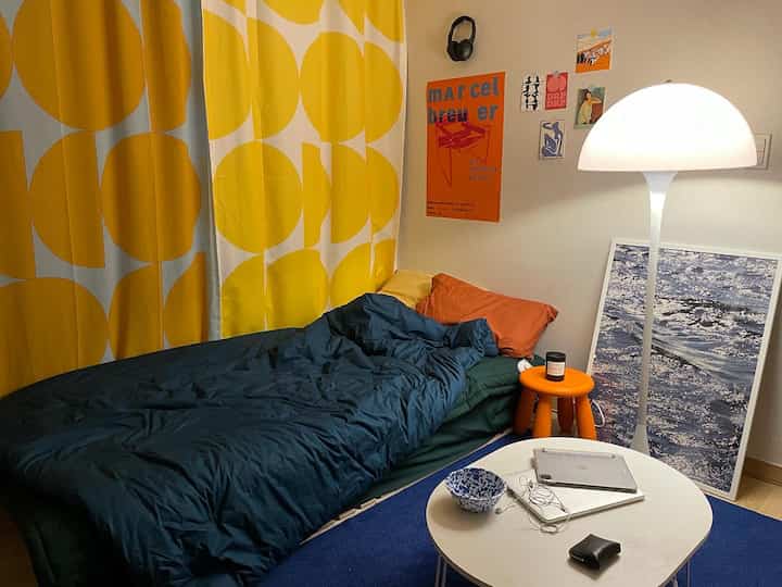 Bedroom with yellow circular patterned curtain and dark blue bedding, white table and orange stool arranged in vivid atmosphere