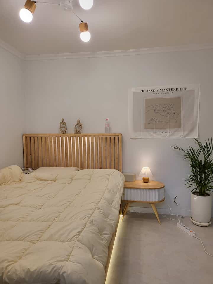 A soft natural color wood tone bedroom featuring a wooden bed, nightstand, fabric poster, and plant in a simple setting