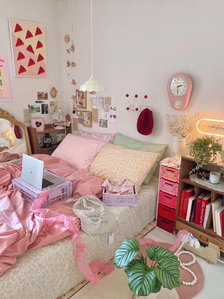 A cozy pink and white bedroom featuring bedding, posters, clock, plants, and organized storage boxes with a stylish atmosphere