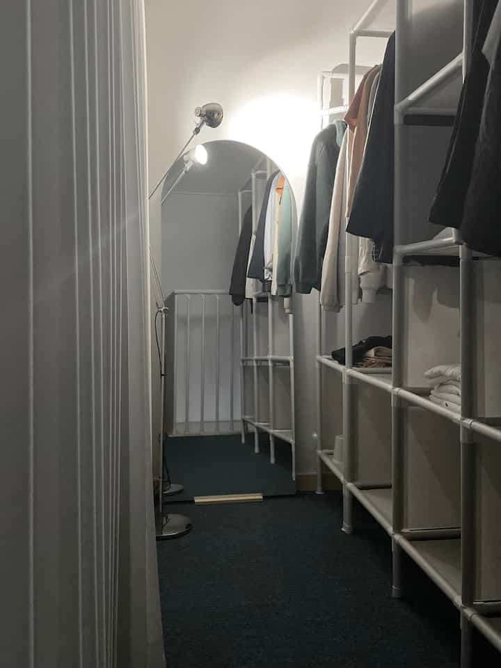 White walls and racks in a narrow, compact walk-in closet featuring an arched full-length mirror and lighting