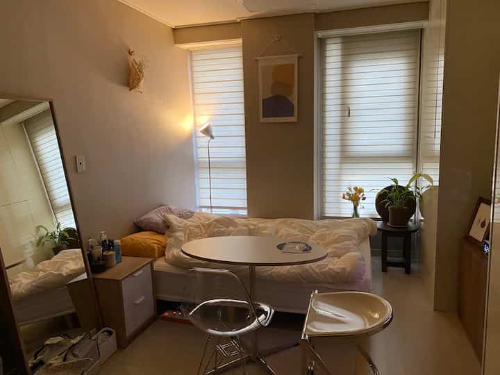 Beige and white toned studio bedroom featuring a bed and small dining table centrally placed with clean, natural decor