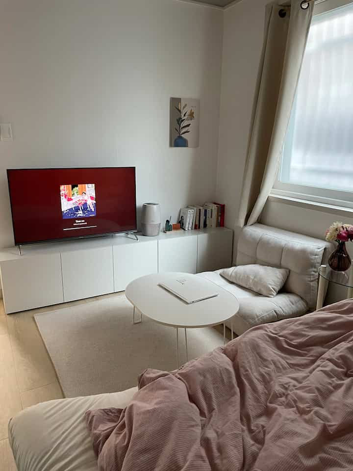 ホワイトとベージュのトーンのワンルーム、コンパクトなソファとコーヒーテーブル、テレビスタンドが配置された自然で落ち着いたインテリア