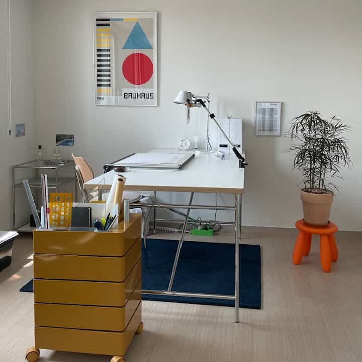A clean white and blue-toned study featuring a large central desk with lamp and an orange stool supporting a potted plant on the right side