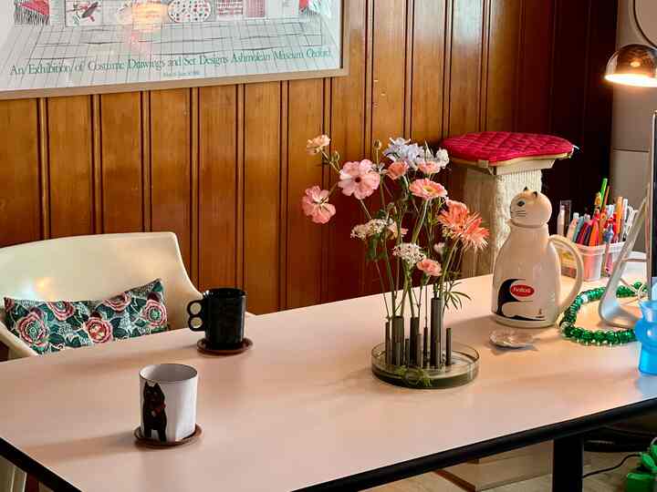 Modern home office space with wood tone wall and white table, featuring a vase of flowers, mugs, and a cat-shaped kettle