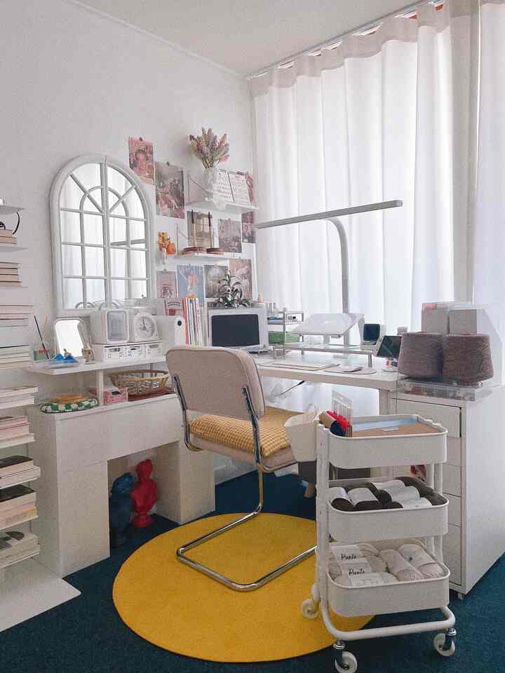 A white and yellow toned craft workspace featuring a large desk, chair, and various storage furniture in a cozy setting