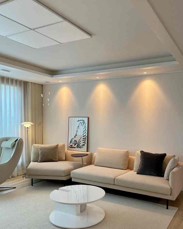 Beige and white toned living room featuring simple modern linear fabric sofa and round coffee table, warm lighting, and minimalist framed artwork creating restrained ambiance