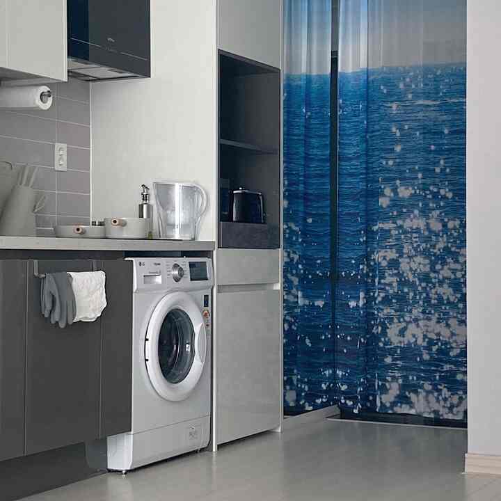 A modern kitchen space in white and gray monotones, featuring a washing machine beside an ocean-themed curtain, with a simple and clean design