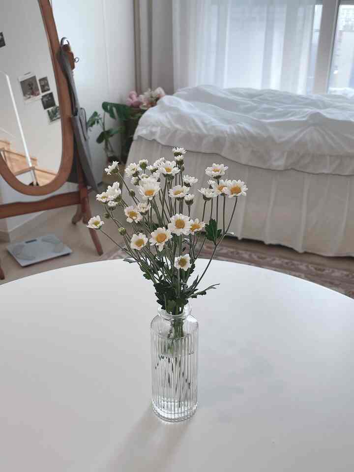 白と木目調のワンルーム寝室、生花の花瓶と木製全身鏡が配置されたナチュラルな雰囲気のインテリア