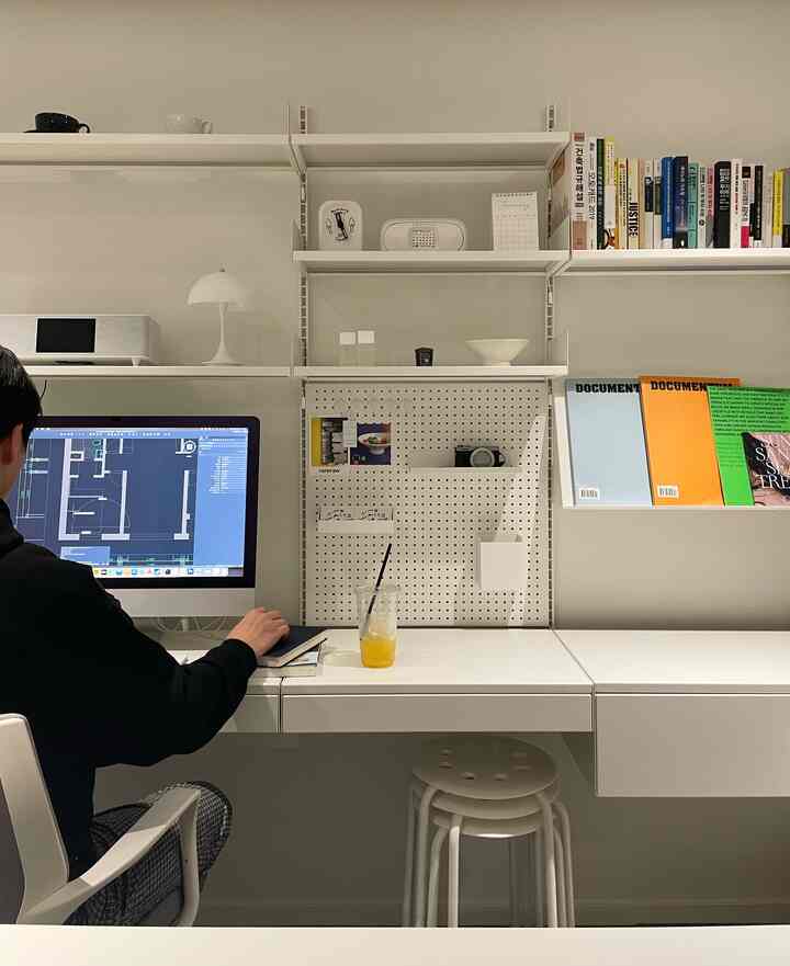 White-toned home office space featuring a spacious desk, shelves, and stools creating a clean modern work environment