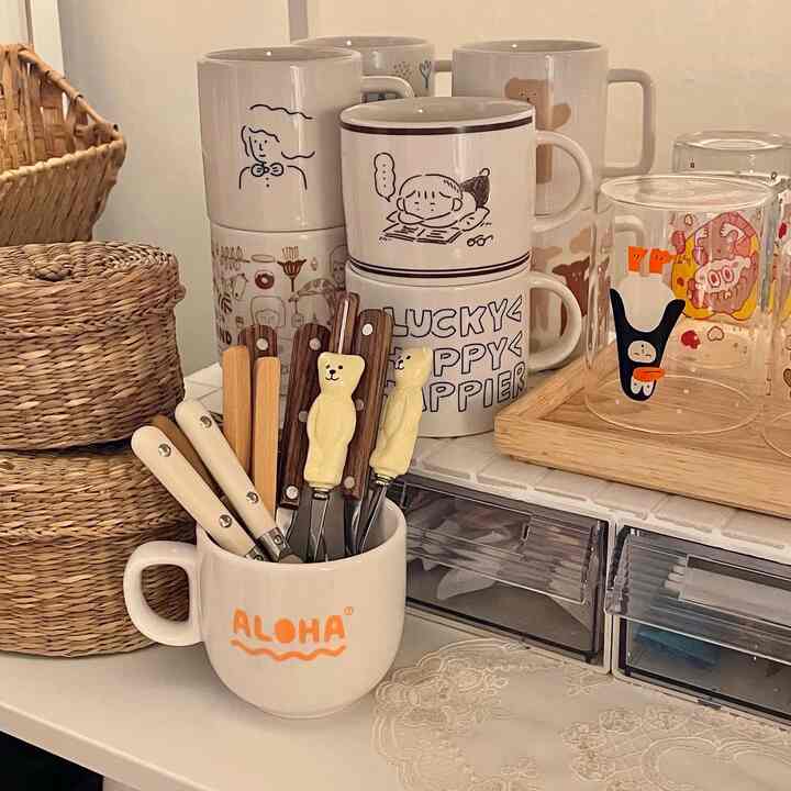 A cozy space featuring natural toned kitchen table with a mug holding wooden handled cutlery and various illustrated mugs arranged