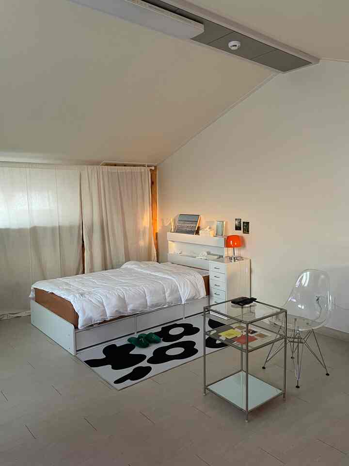 Natural toned bedroom featuring a white bed, nightstand with an orange lamp, and a transparent side table in a clean setting