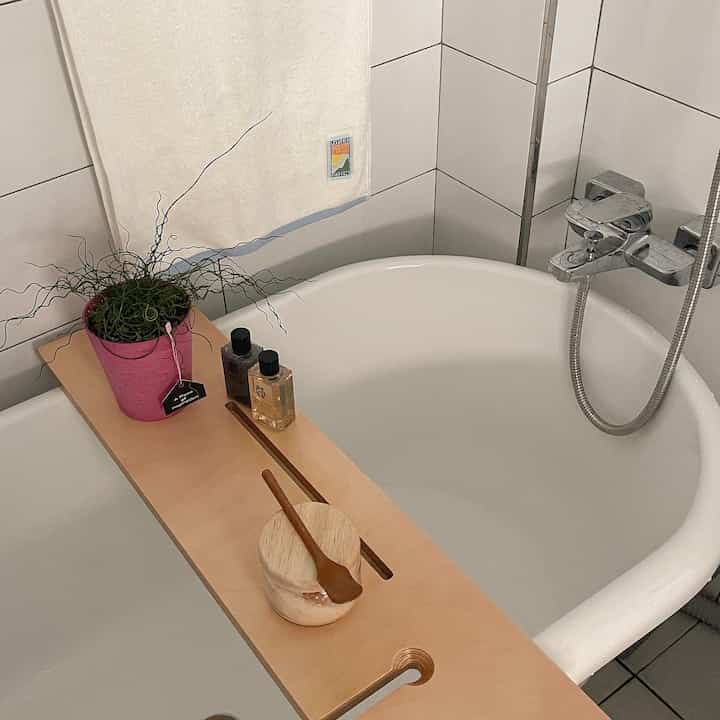 白いタイル壁と浴槽に、木目調のバストレイが植物やバス用品を配した、清潔感のあるバスルーム