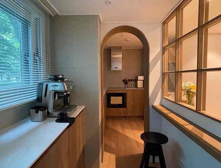 ウッドトーンとベージュが主体のアーチ型入り口のあるキッチンと、左側の窓辺にコーヒーマシンが置かれた落ち着いたおうちカフェ空間