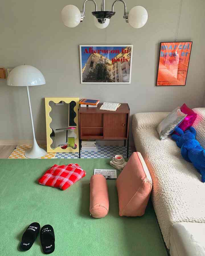 A living room with beige walls and green rug featuring a cream sofa, wooden console, floor lamp, and framed posters in a mid-century modern style