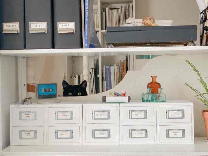White shelves and storage drawers organized in a workspace featuring a black cat peeking over the shelf, cozy atmosphere