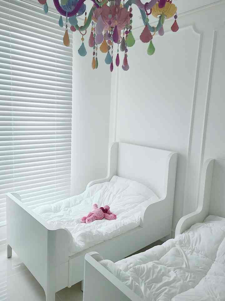 White-toned kids' room featuring two white beds, white blinds, and a colorful chandelier in a clean setting