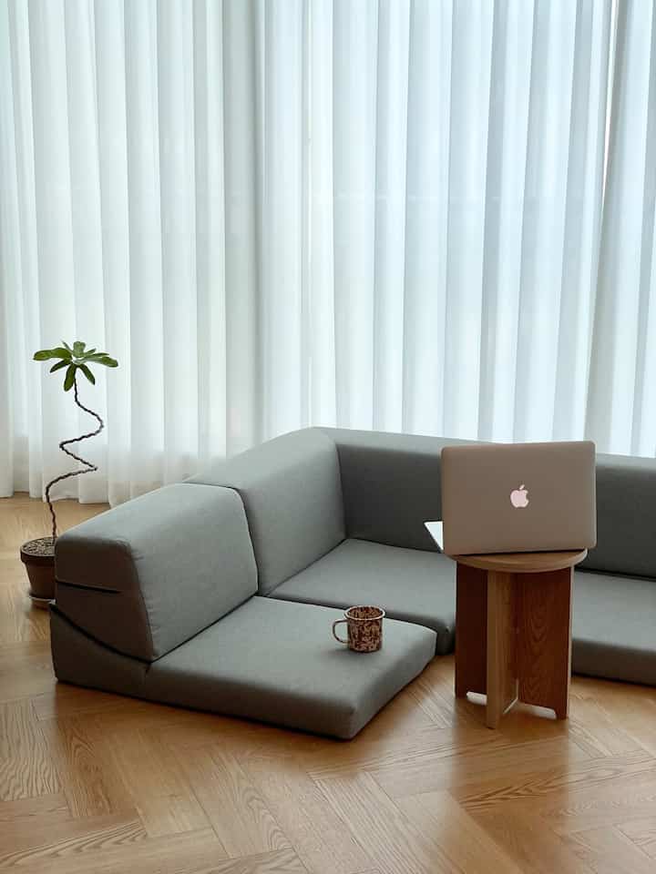 ライトグレーのモジュラー式座椅子ソファと木製サイドテーブル、明るいカーテンが配された自然なリビング空間