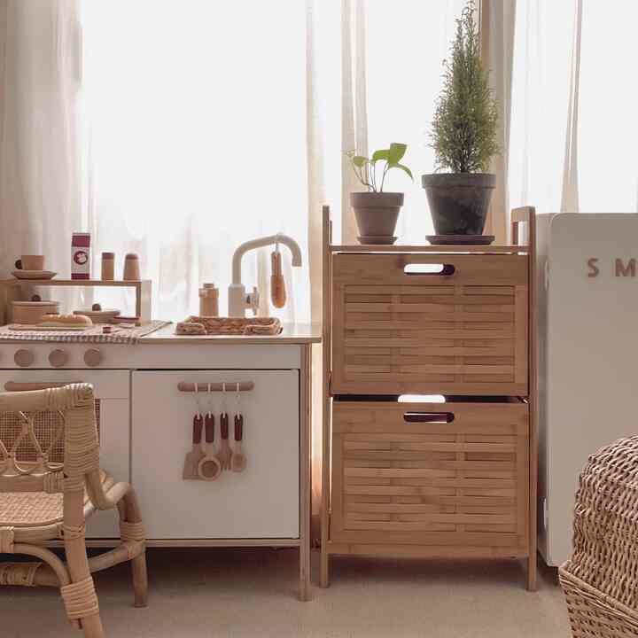 Natural-toned kitchen space featuring a play kitchen and bamboo storage cabinet, complemented by soft beige curtains and carpet creating a cozy atmosphere