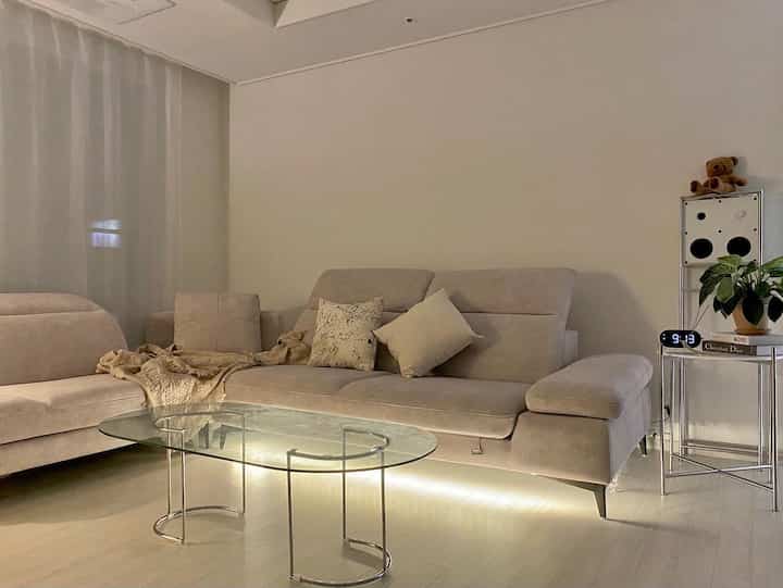 Beige and white toned living room featuring beige sectional sofa and transparent glass coffee table in a natural modern style space