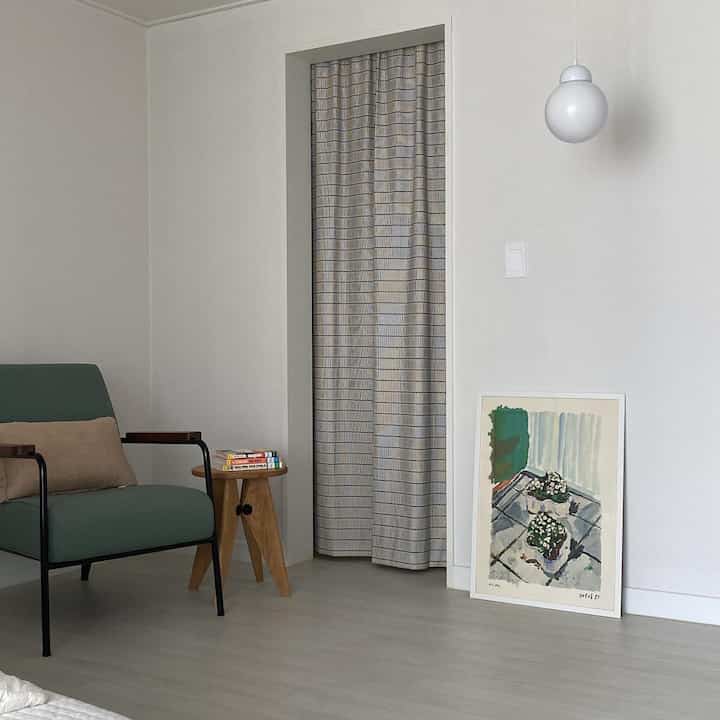 白とナチュラルな色調のリビング、グリーンのアームチェアと木製スツール、ドアカーテンと額装アートが配置されたシンプルモダンなインテリア