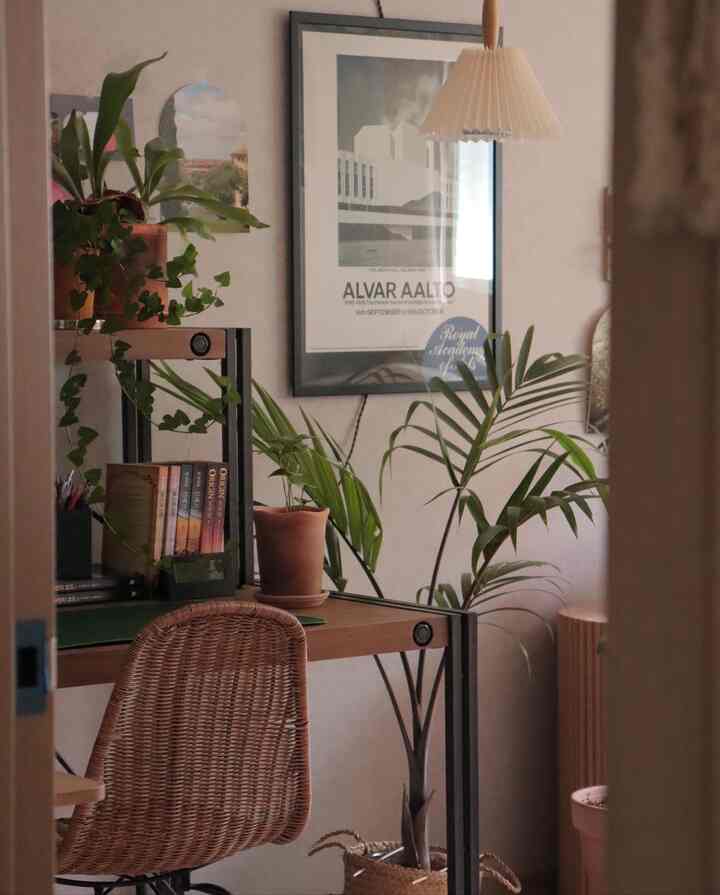 Natural wood-toned bedroom home office with rattan chair, abundant plants, wall art, and pendant light creating a cozy atmosphere