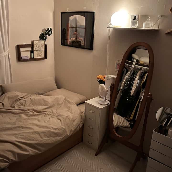 A small bedroom with white walls and wood-toned furniture, featuring a full-length mirror and dresser creating a cozy atmosphere