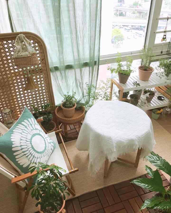 White tablecloth, natural wood-tone furniture, green curtains, and various plants creating a cozy veranda space