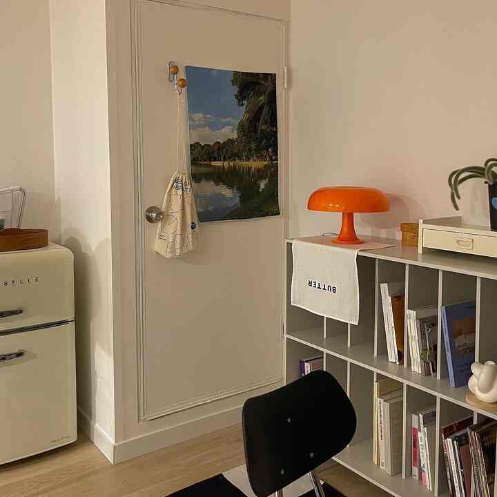 白い壁と明るい木目調の床のワンルーム、オレンジ色のテーブルランプと収納棚が配置されたミニマルでレトロなリビング空間