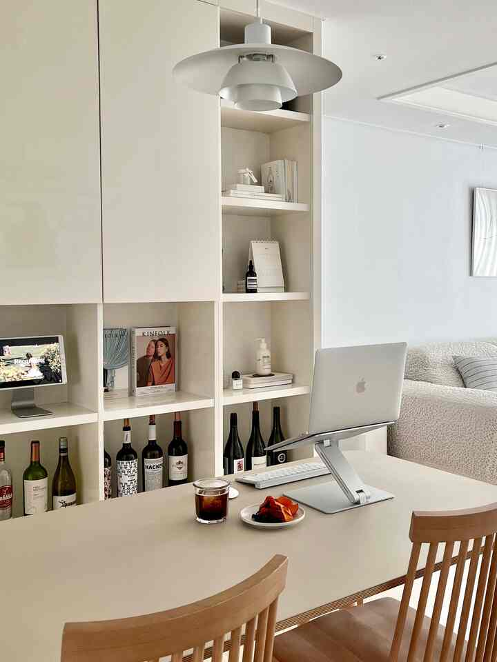 A white and beige tone home office space featuring a clean bookshelf, laptop stand on dining table, and wooden chairs, creating a modern cozy atmosphere