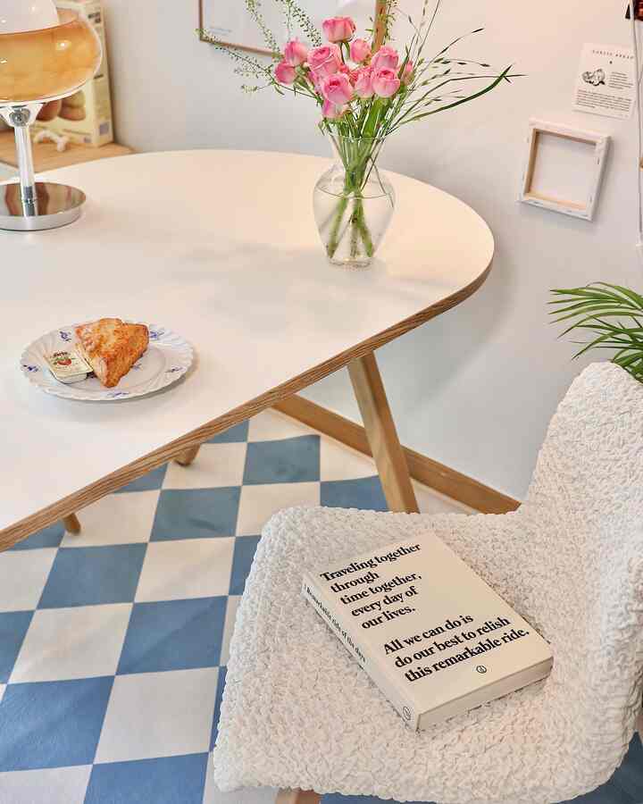 White and natural color toned living room featuring an oval dining table, fluffy chair covers, transparent vase, and checkered rug creating a cozy interior