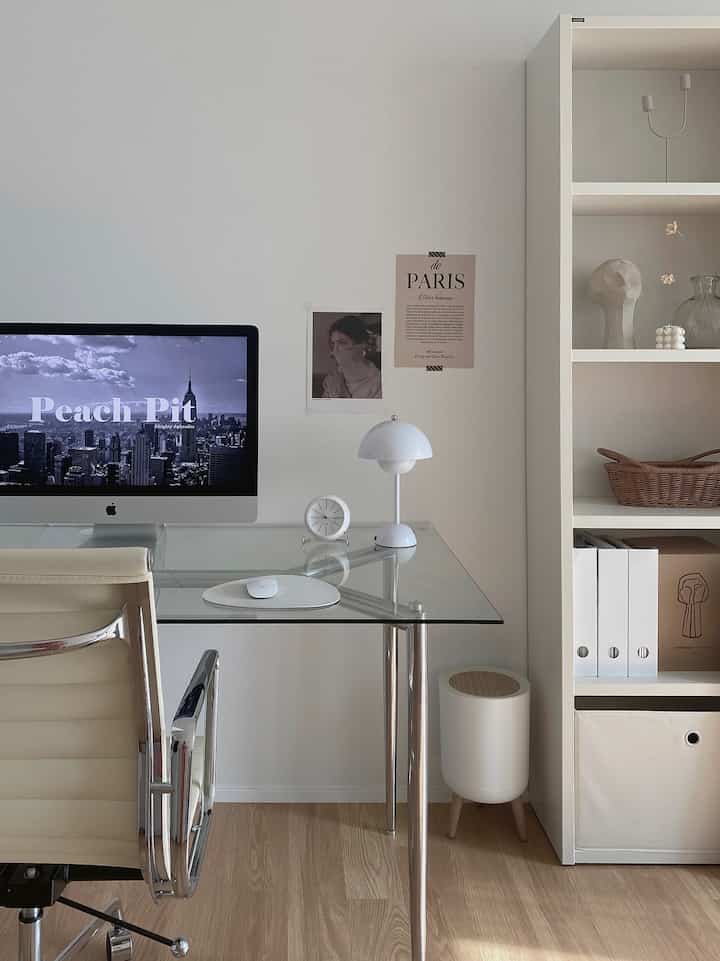 A modern study with white and wood tones featuring a glass desk, office chair, organized bookshelf, and simple decor elements