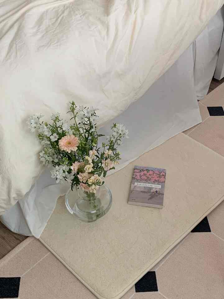 Beige and white toned bedroom space featuring cream duvet and white bed skirt, cozy atmosphere with transparent vase holding flowers on a beige bath mat
