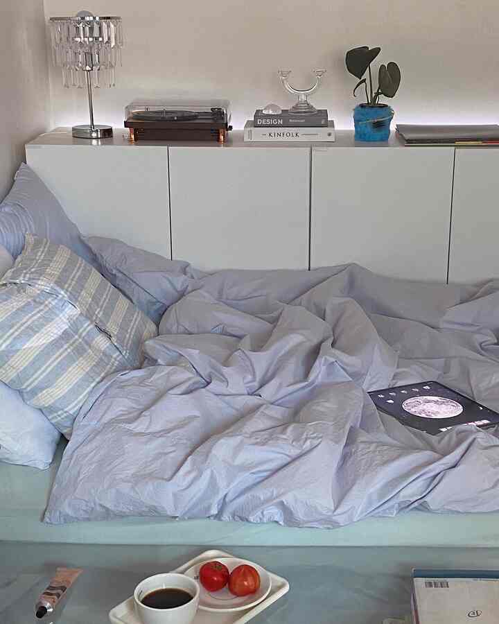 Modern bedroom with white and blue tones featuring tidy bedding, books, and a plant on a sleek cabinet