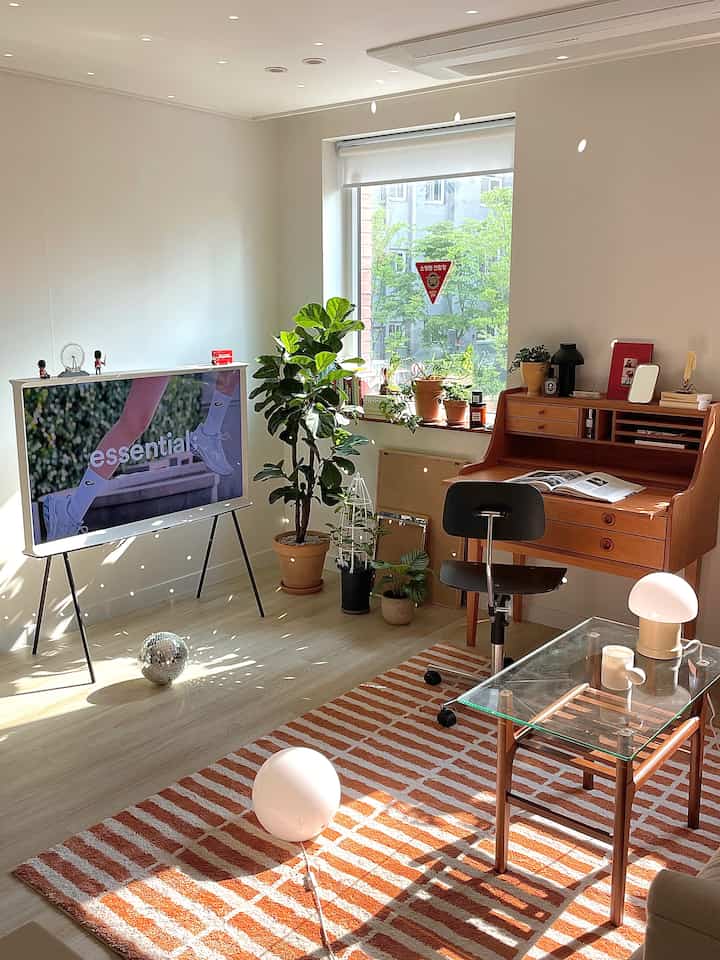 A white and brown toned living room featuring plants by the window, a desk, and a rug creating a natural modern atmosphere