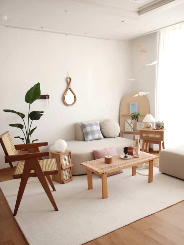 Natural beige and white toned living room featuring rattan table and armchair with tall green plant, creating a simple and cozy atmosphere