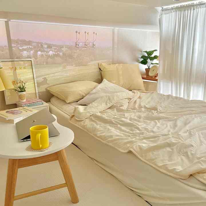A beige and wood-tone bedroom featuring a bed, desk with laptop, curtains, and plants creating a cozy natural atmosphere
