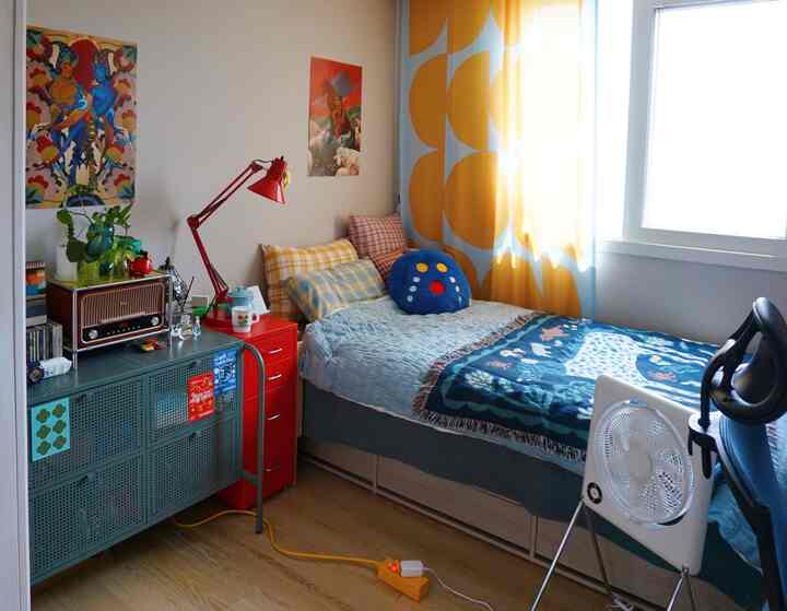 Compact bedroom with bright yellow patterned curtains and blue bedspread, featuring red dresser and desk
