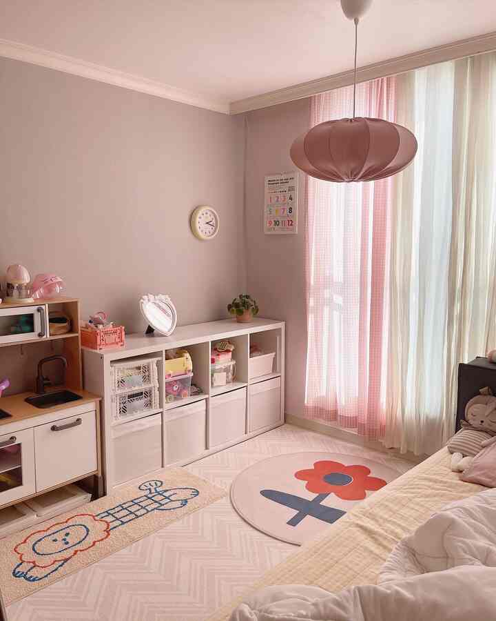 パステルピンクとホワイトの子供部屋、本棚、ベッド、ラグ、時計とカレンダーが配置された雰囲気のあるインテリア