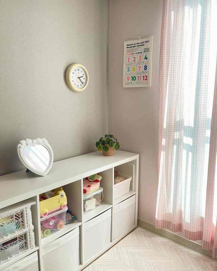 ピンク色調の子供部屋、中央に白い収納棚、壁に時計とカレンダーが掛かり、ピンクのギンガムカーテンが明るい雰囲気を演出しているインテリア