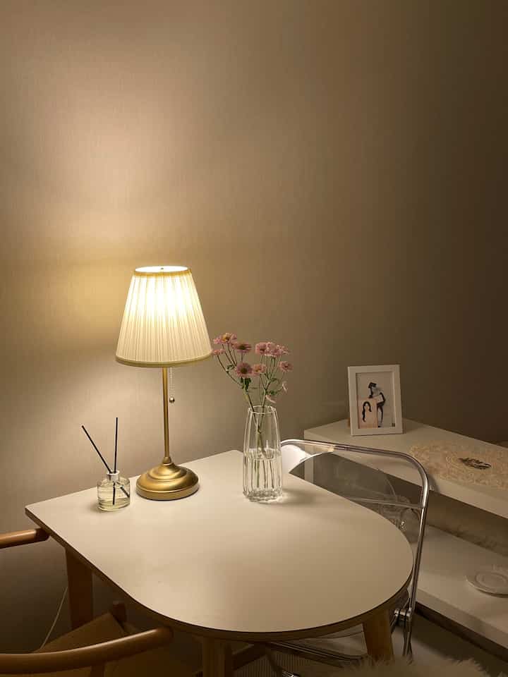温かみのあるゴールド照明と白いテーブル、花瓶と透明な椅子が配置されたシンプルでモダンな一人暮らしの空間