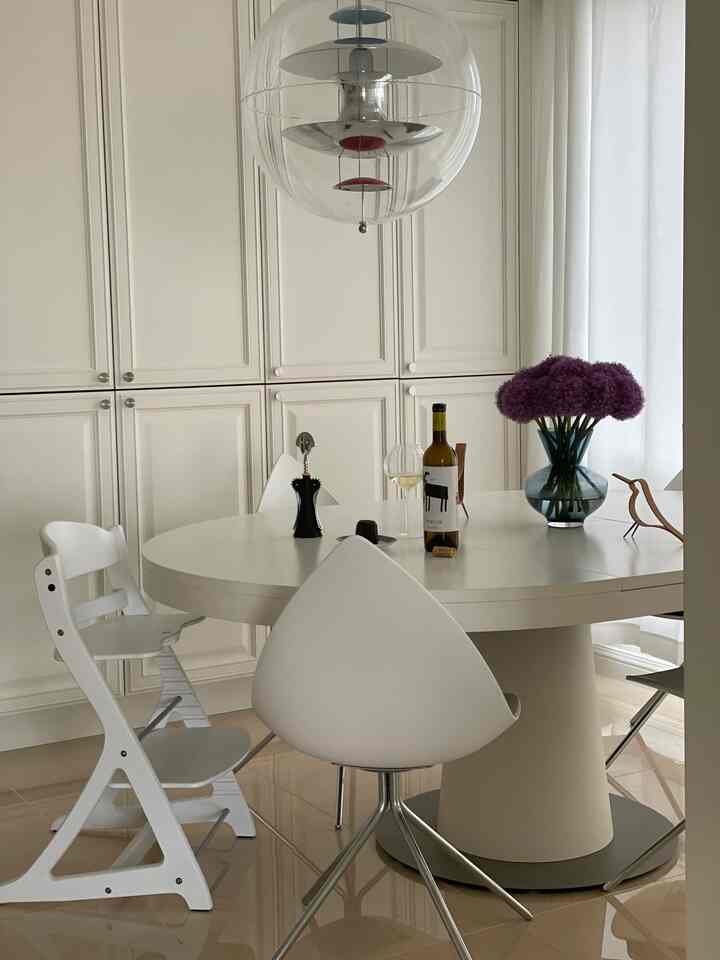 White and natural-toned dining room featuring round dining table, baby chair, wine glass, and flower vase in a warm setting
